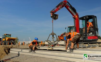 Mit Hilfe eines Baggers, der die neue Betontreppe an einer Kette trägt, wird dieses Teil von zwei Mitarbeitern Millimeter genau an die finale Position gesetzt.