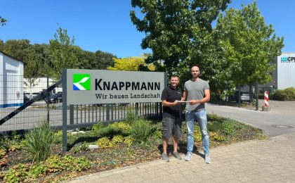 Christopher Kuhl und Thassilo Thols stehen vor dem Unternehmensschild. Es wird ein Geschenk zum bestandenen Bachelor überreicht.